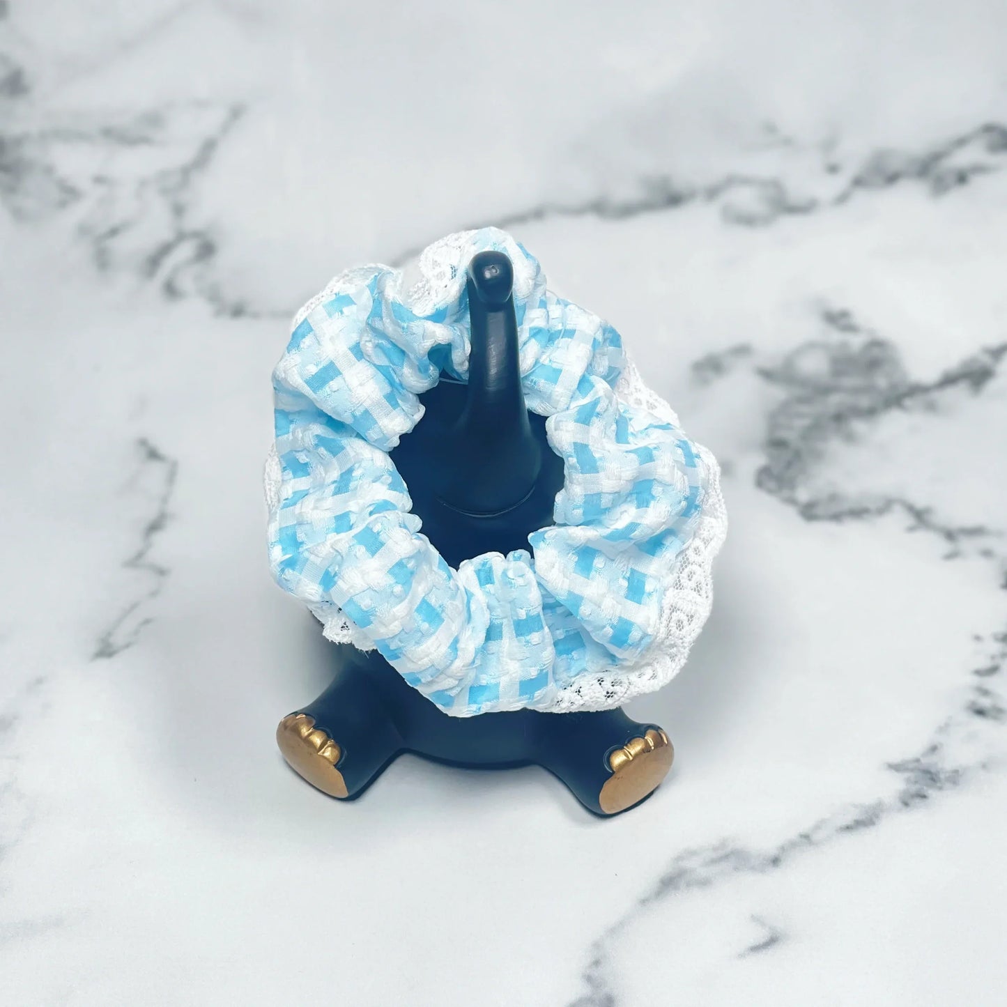 Blue and white check scrunchies with lace displayed on black ring holder on marble background