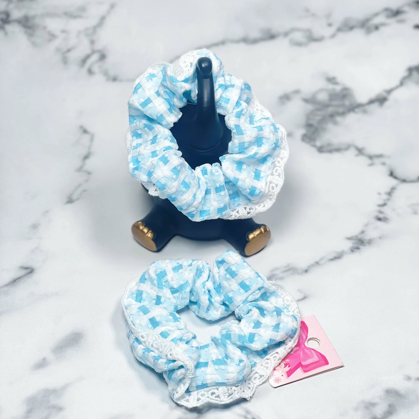 Blue and white check scrunchies with lace detail displayed on marble surface and black stand