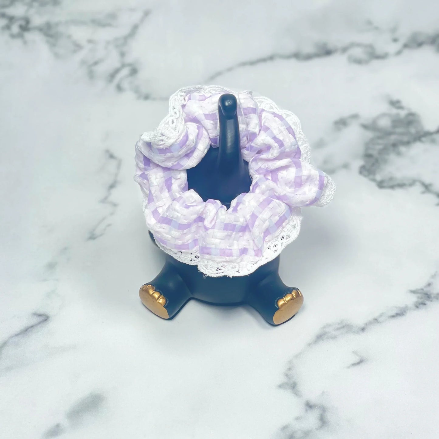 Purple and white check scrunchies with lace displayed on a black stand with gold details on marble surface