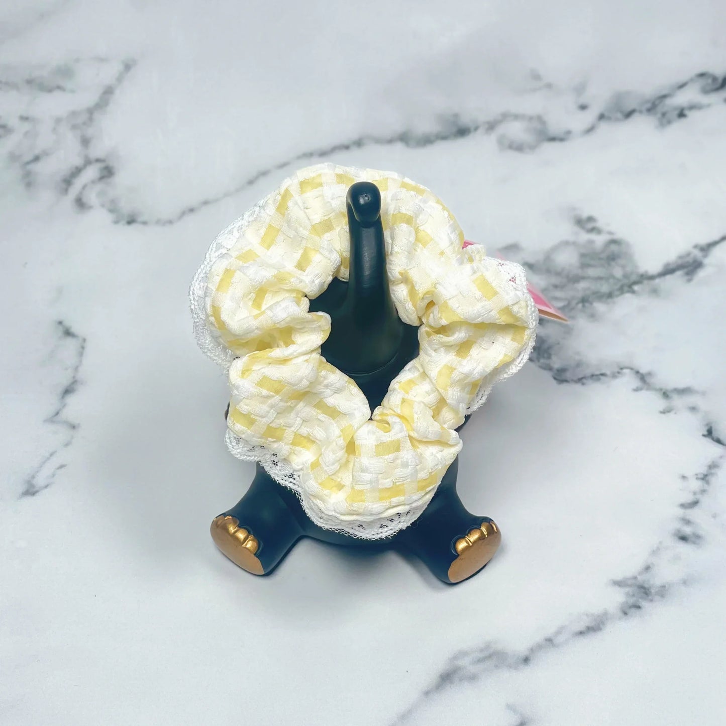 Yellow and white check scrunchies with lace displayed on a black stand on a marble surface