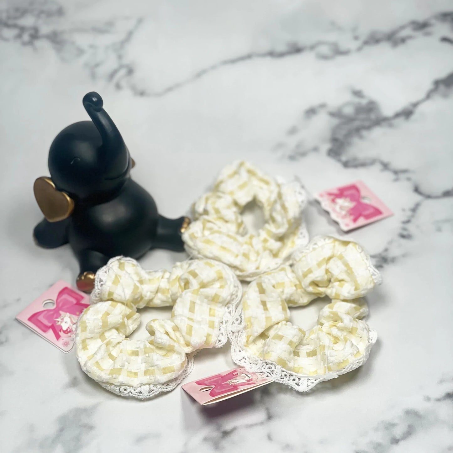 Set of yellow and white check scrunchies with lace trim displayed on marble surface next to black elephant figurine