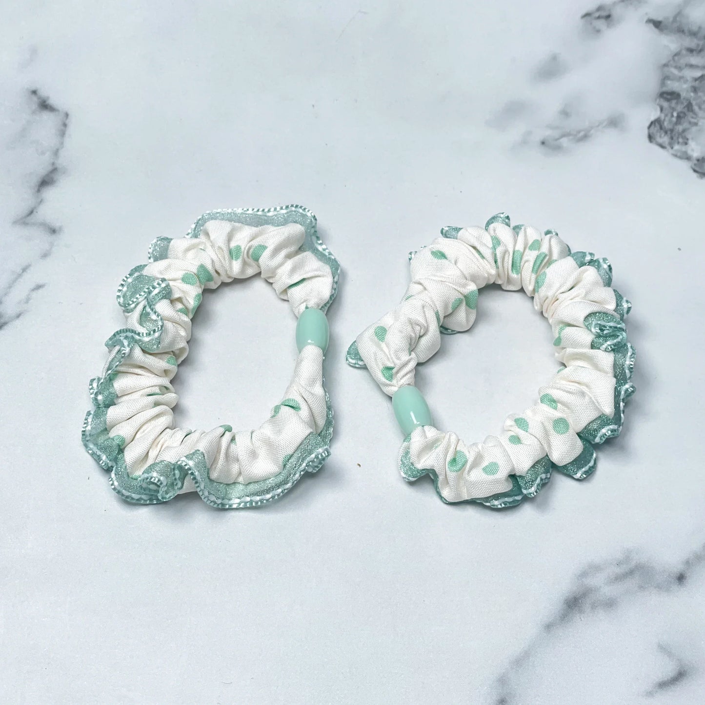 Two white and green frill polka dot hair ties with ruffled edges on a marble background