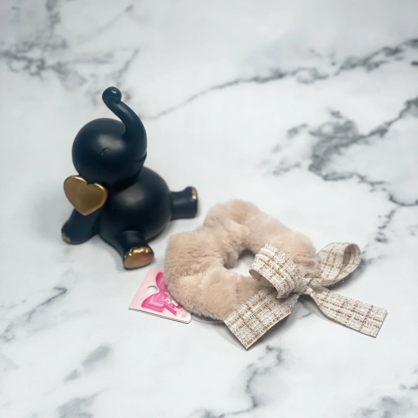 Beige fur scrunchie with check ribbon on marble surface next to black elephant figurine