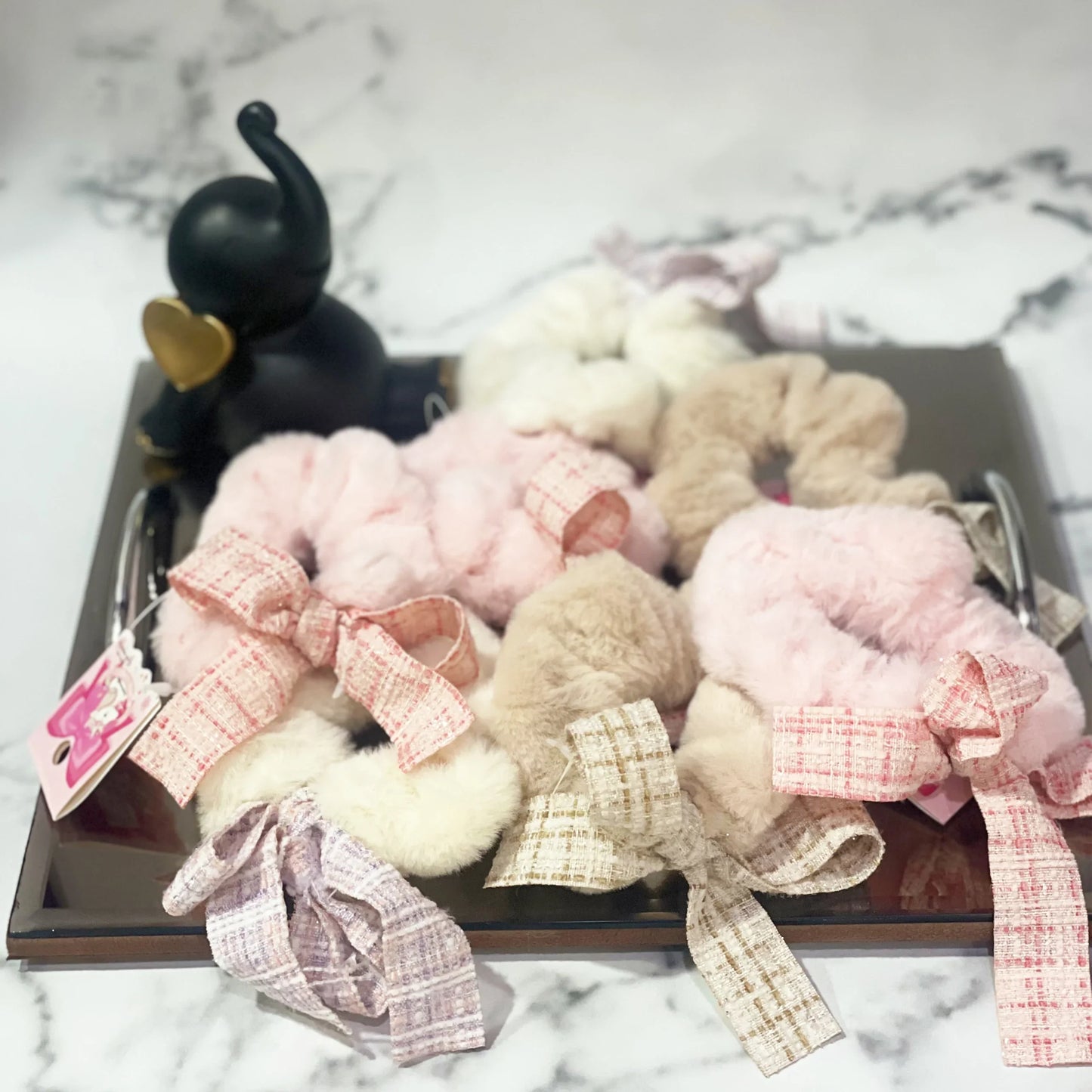 Assorted soft fur scrunchie with check ribbon in pink, beige, and lavender on a black tray