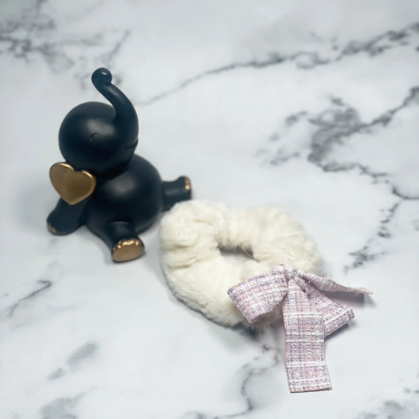 White fur scrunchie with check ribbon laid on marble surface next to black elephant figurine