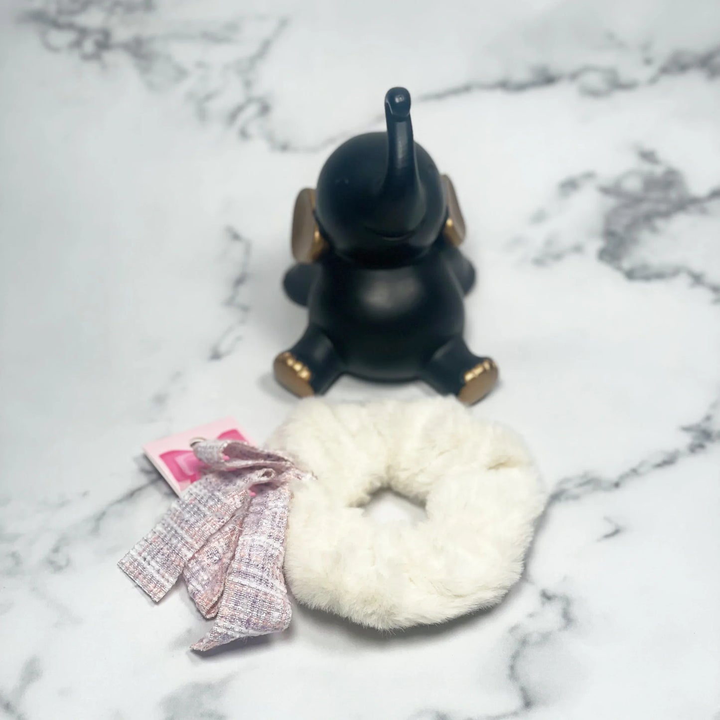 Fur scrunchie with check ribbon in soft white placed on marble surface next to decorative black elephant