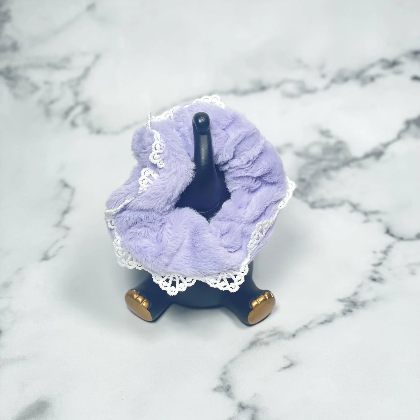 Lavender fur scrunchies with lace trim displayed on a black stand over a marble surface