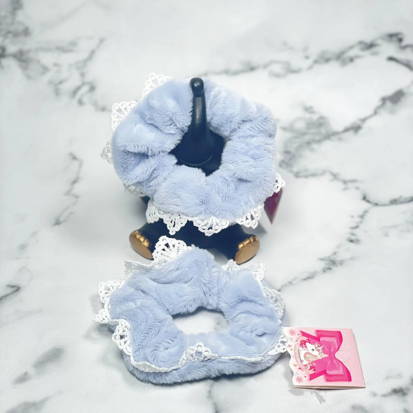 Light blue fur scrunchies with lace trim displayed on a marble surface with a pink tag