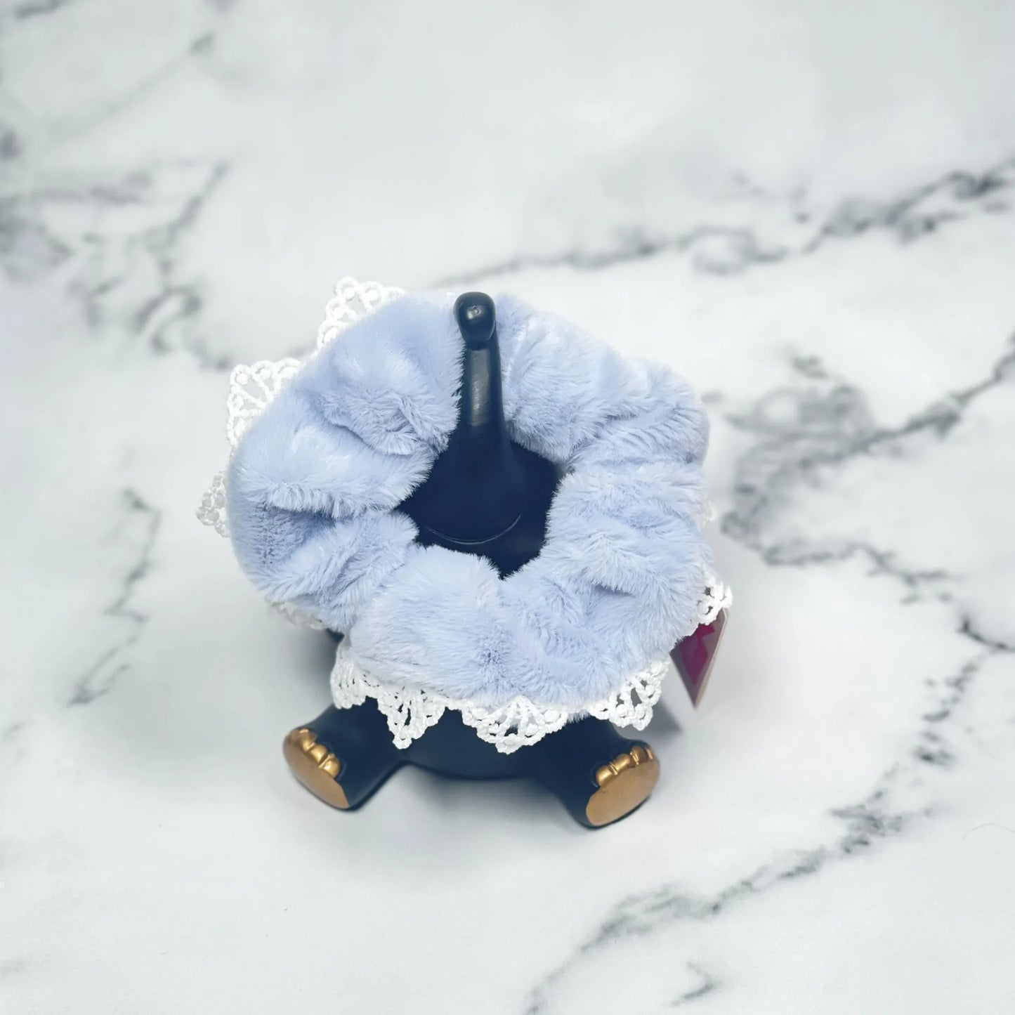 Light blue fur scrunchies with lace trim displayed on a black stand on marble background