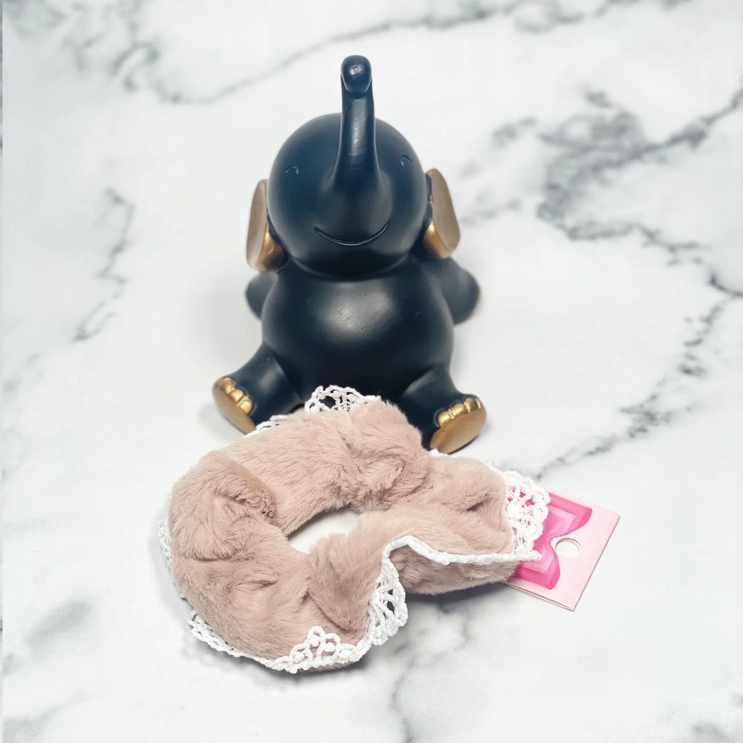 Beige fur scrunchies with lace detail displayed on marble surface beside a black decorative elephant