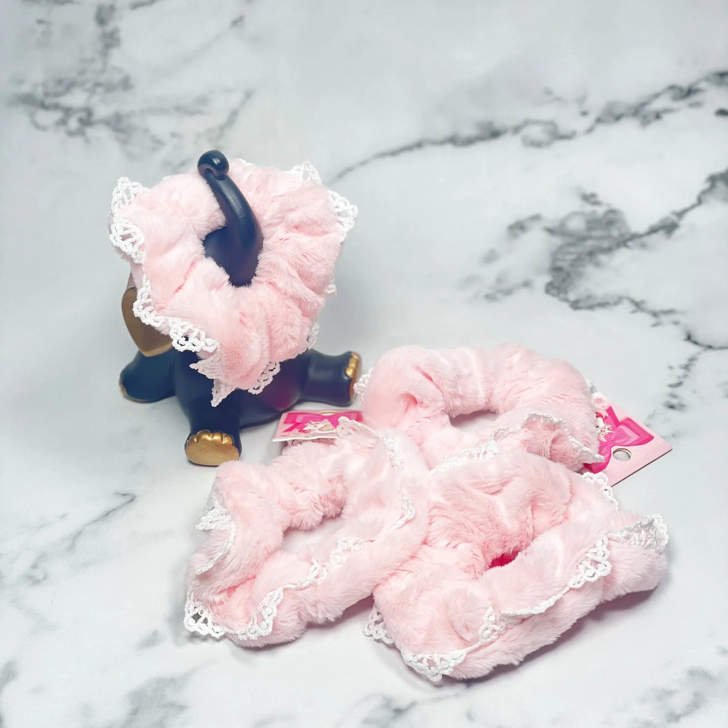 Soft pink fur scrunchies with lace trim displayed on marble surface with decorative elephant