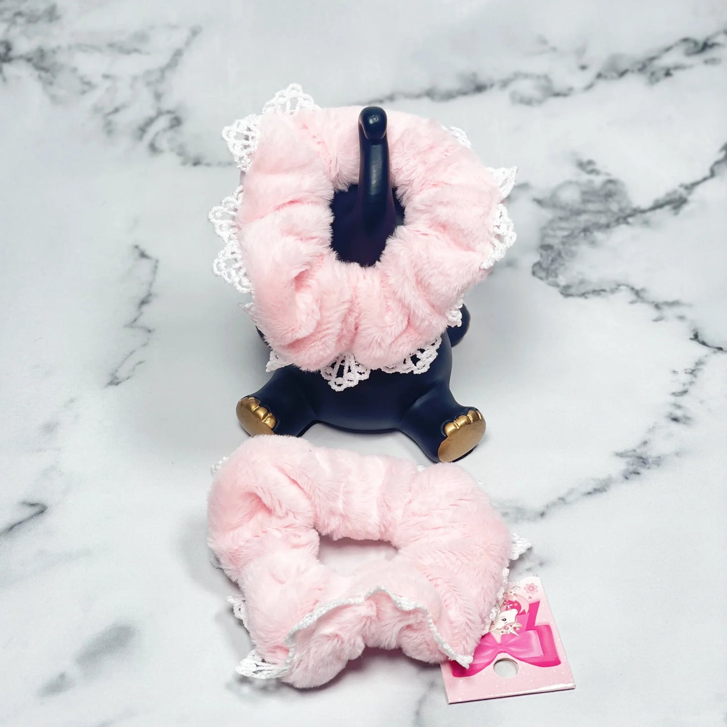 Soft pink fur scrunchies with lace trim, displayed on a marble surface and black stand