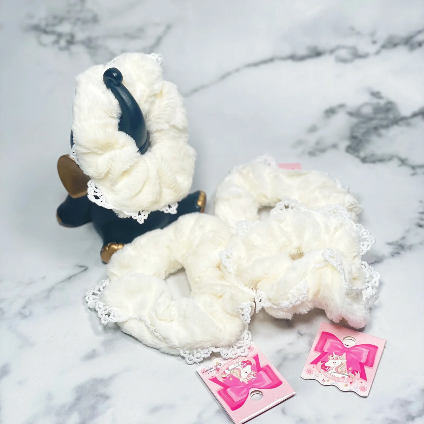 White fur scrunchies with lace edges displayed on a marble surface with pink tags
