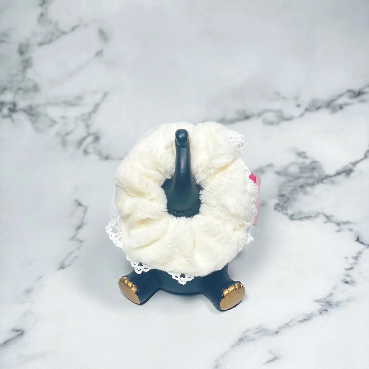 White fur scrunchies with lace detail displayed on a black stand with gold feet on marble surface