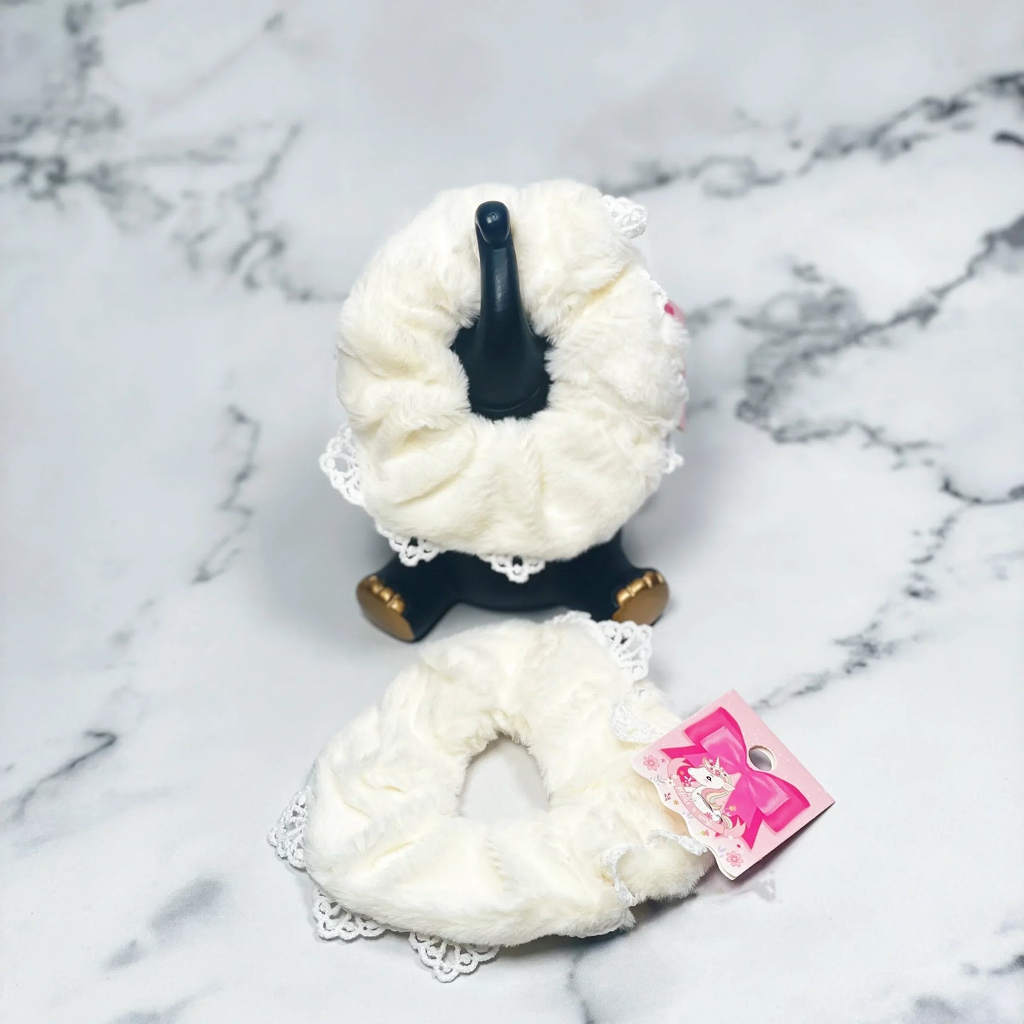White fur scrunchies with lace trim displayed on black stand and marble surface
