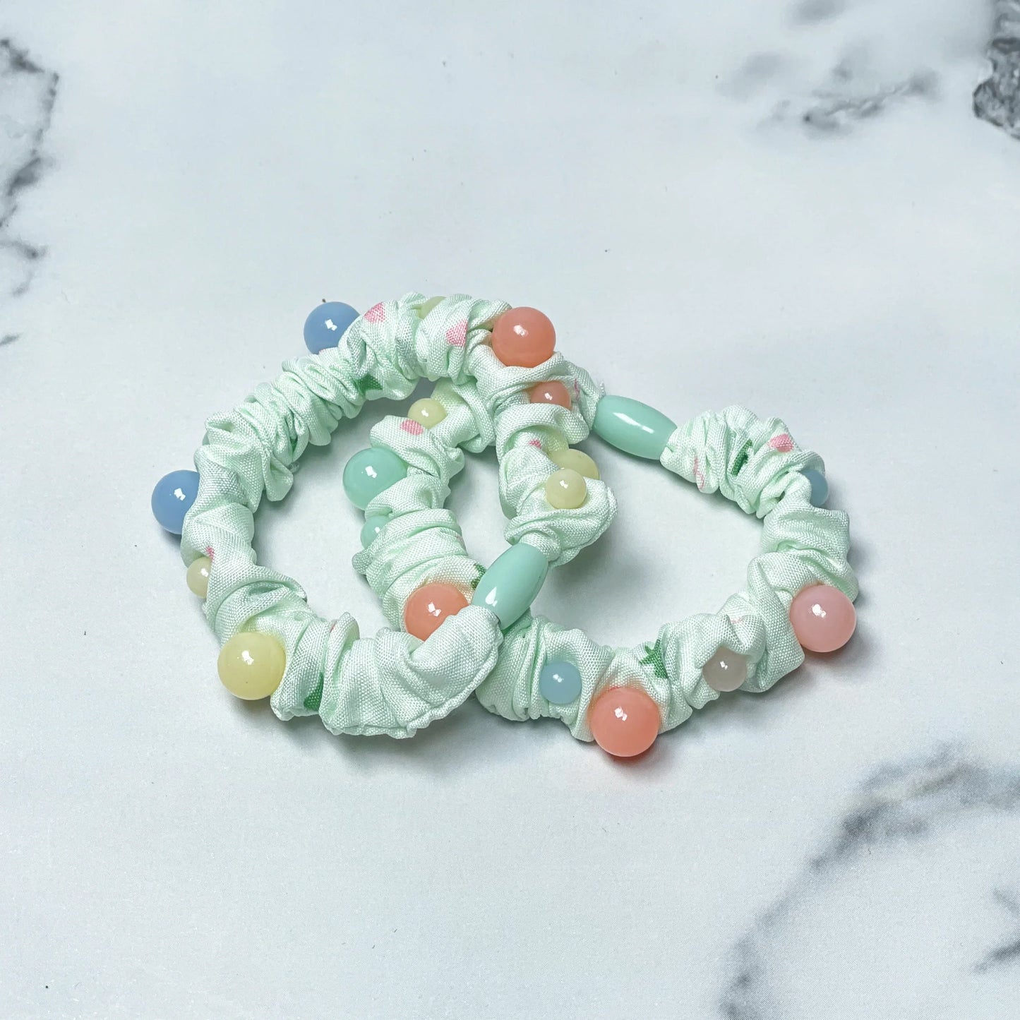Light green printed hair ties with colorful bead accents on white marble surface