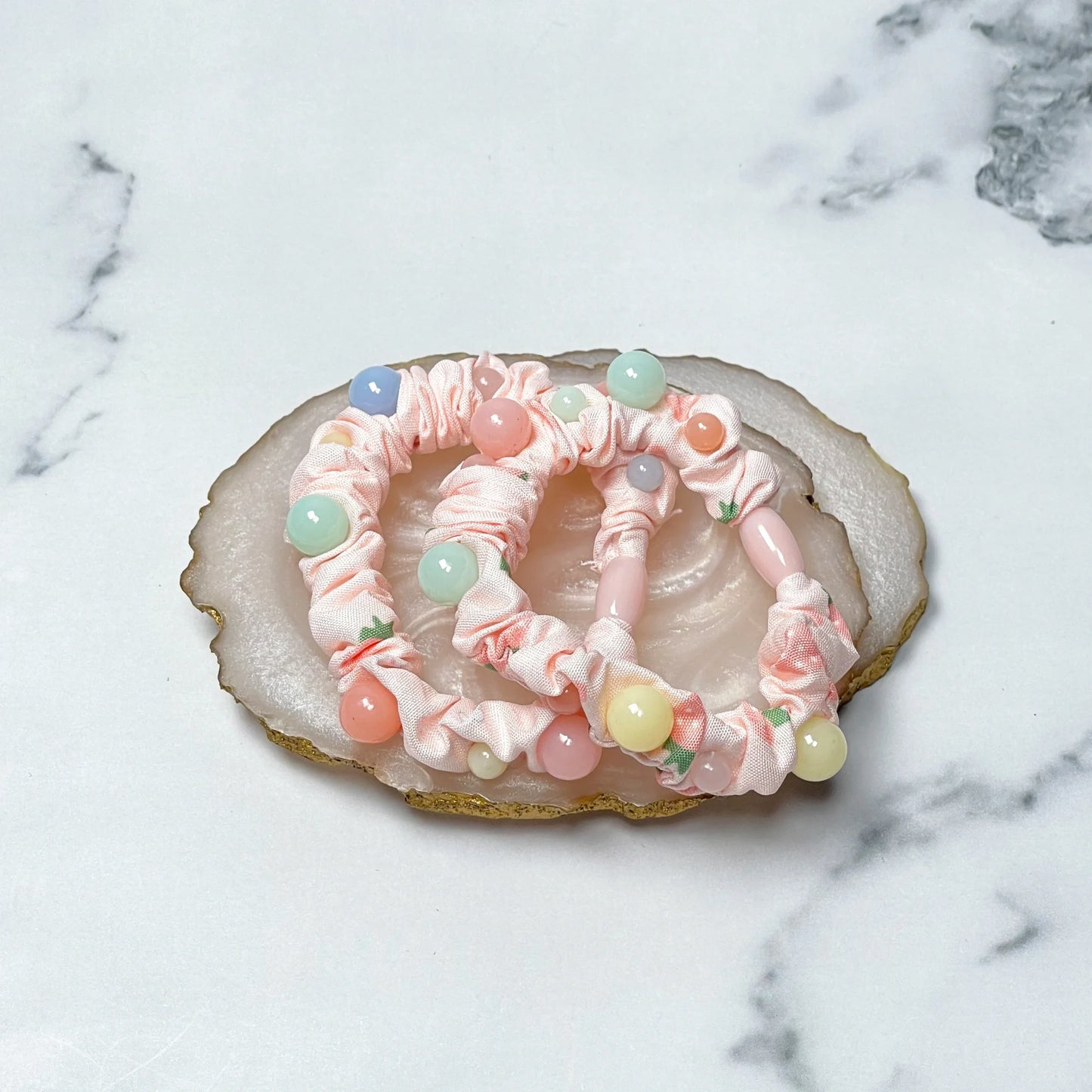 Pink scrunchie printed hair ties with colorful beads displayed on a marble and agate stone surface