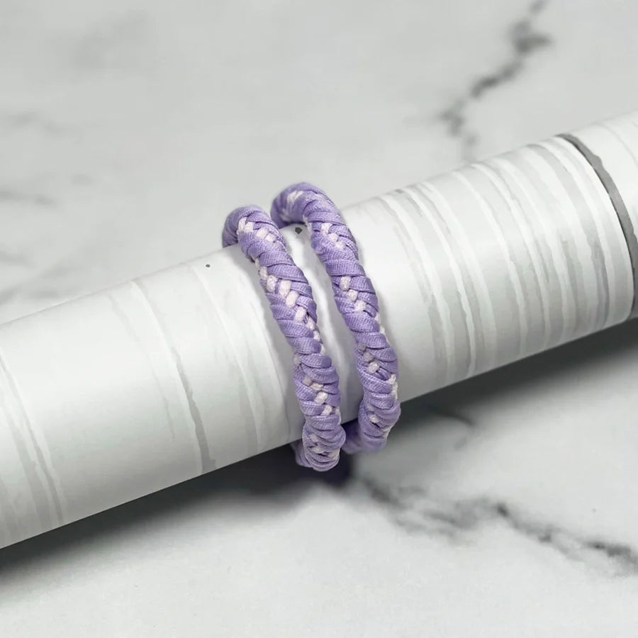Two purple braided thick hair ties displayed on a white cylindrical holder on a marble surface