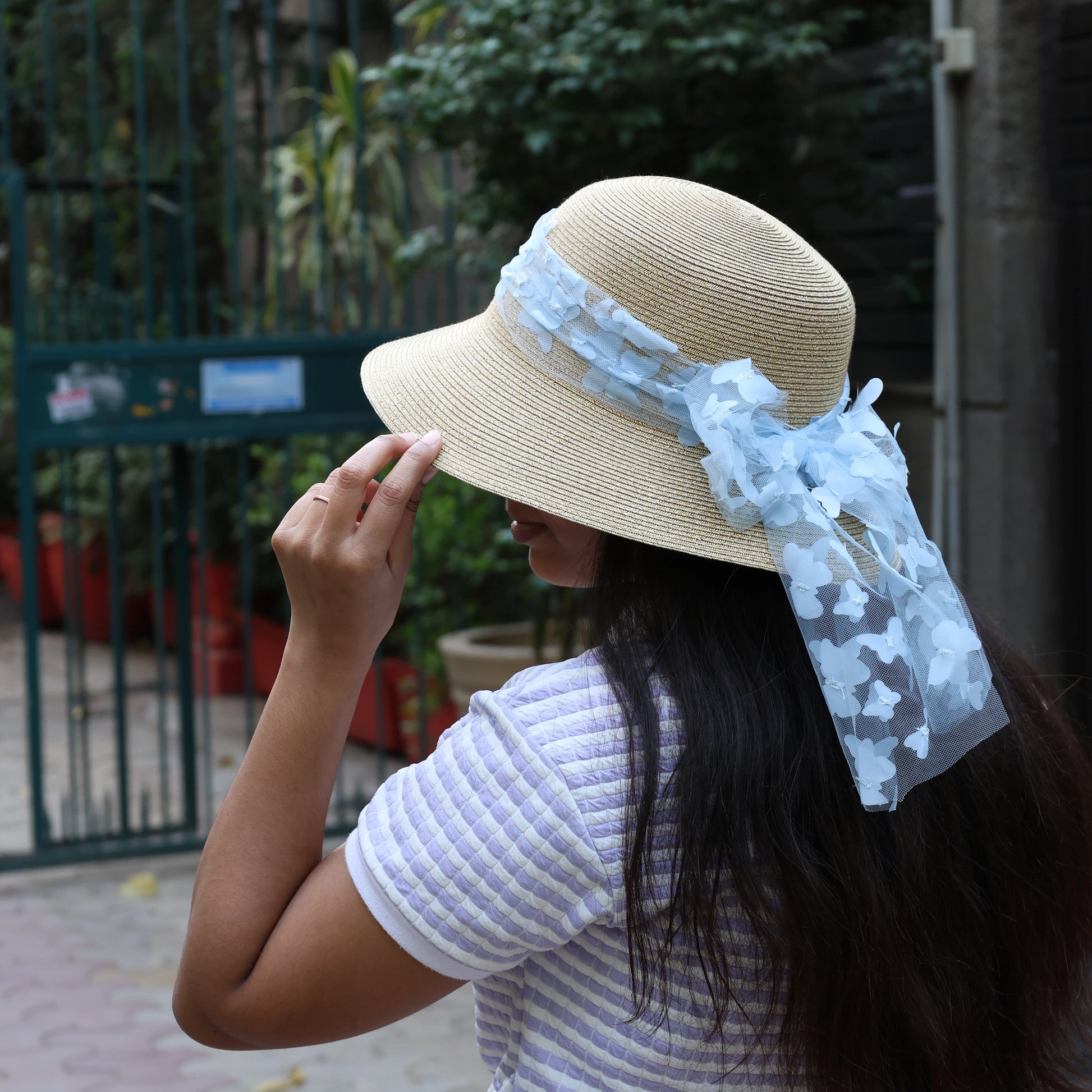 Beige straw round hat with blue ribbon worn by woman with long dark hair in outdoor setting