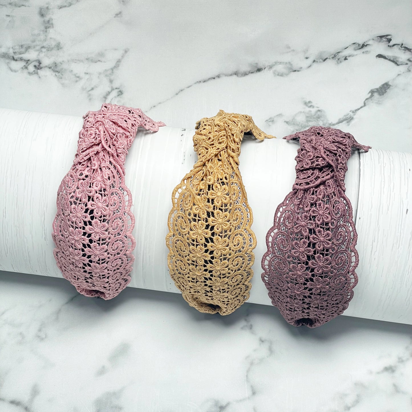 Three Broad Lacy Knotted Hairbands in pink beige and mauve displayed on a white cylindrical stand