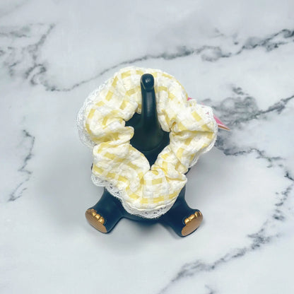 Yellow and white check scrunchies with lace displayed on a black stand on a marble surface