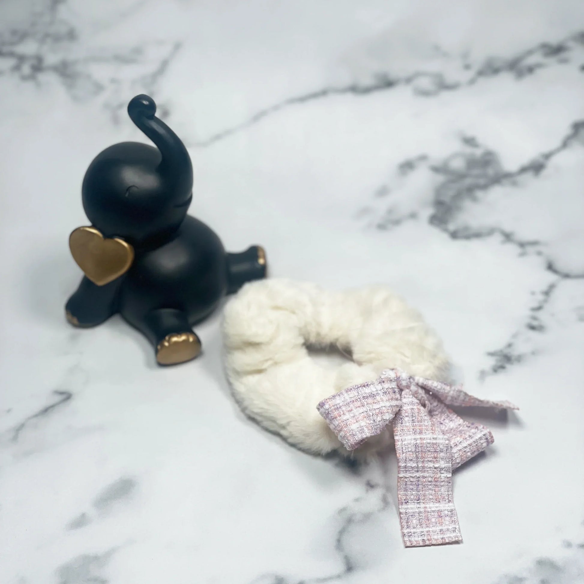 White fur scrunchie with check ribbon laid on marble surface next to black elephant figurine