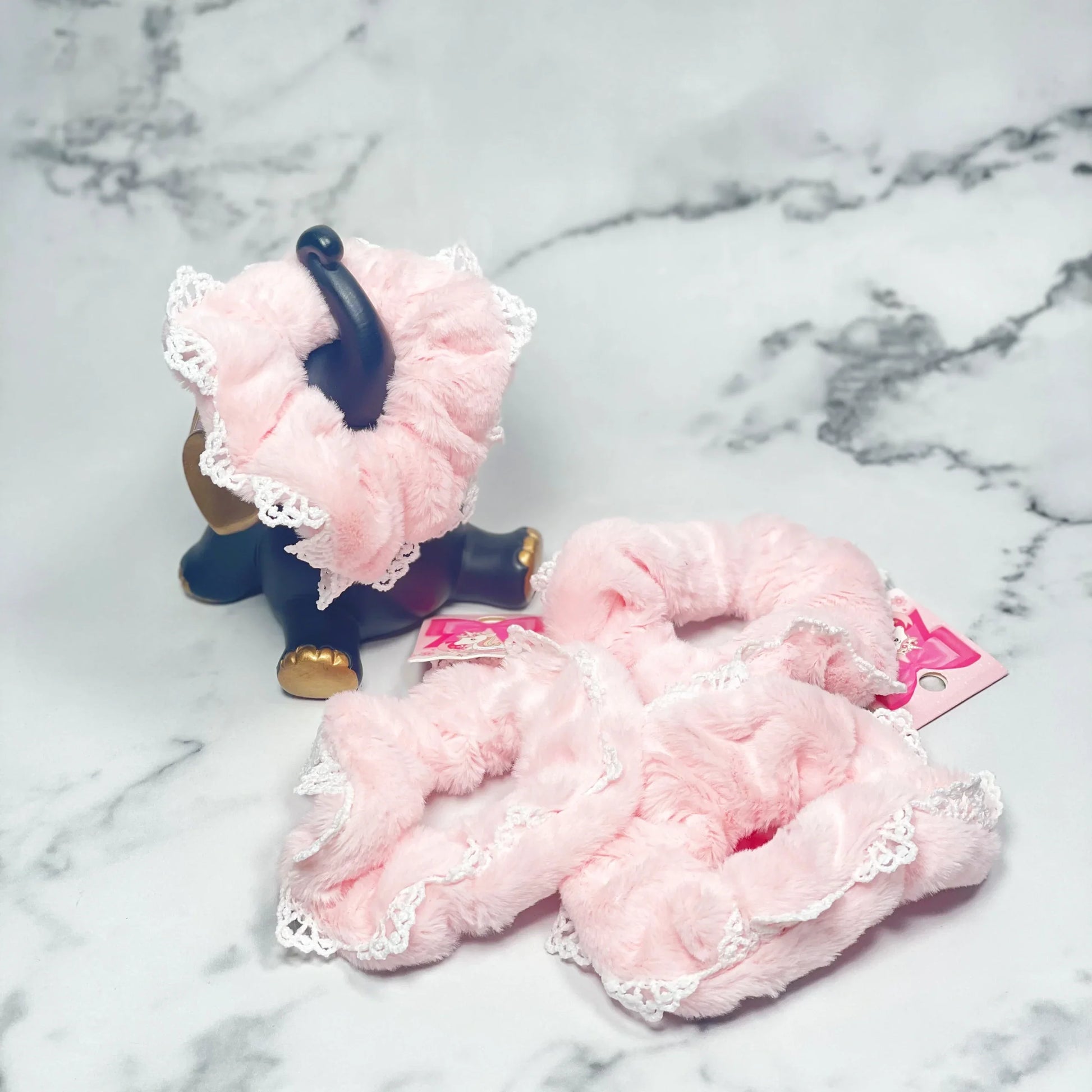 Soft pink fur scrunchies with lace trim displayed on marble surface with decorative elephant