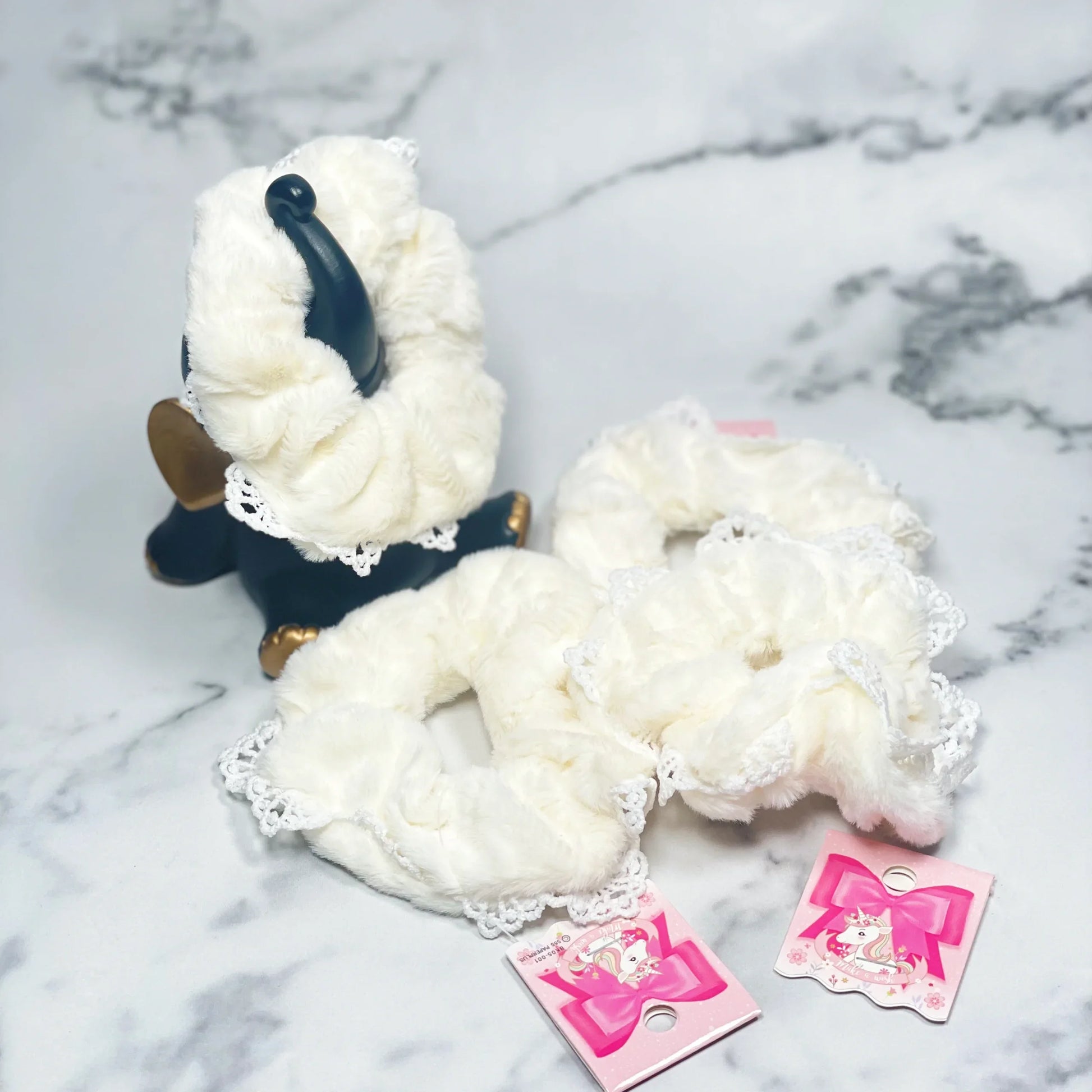 White fur scrunchies with lace edges displayed on a marble surface with pink tags