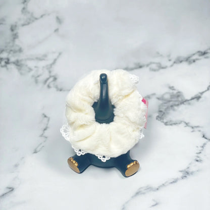 White fur scrunchies with lace detail displayed on a black stand with gold feet on marble surface