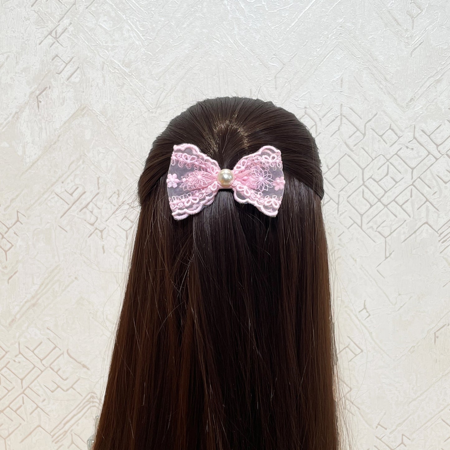 Pink lacy alligator bow clips with pearl center worn on dark brown hair against textured background