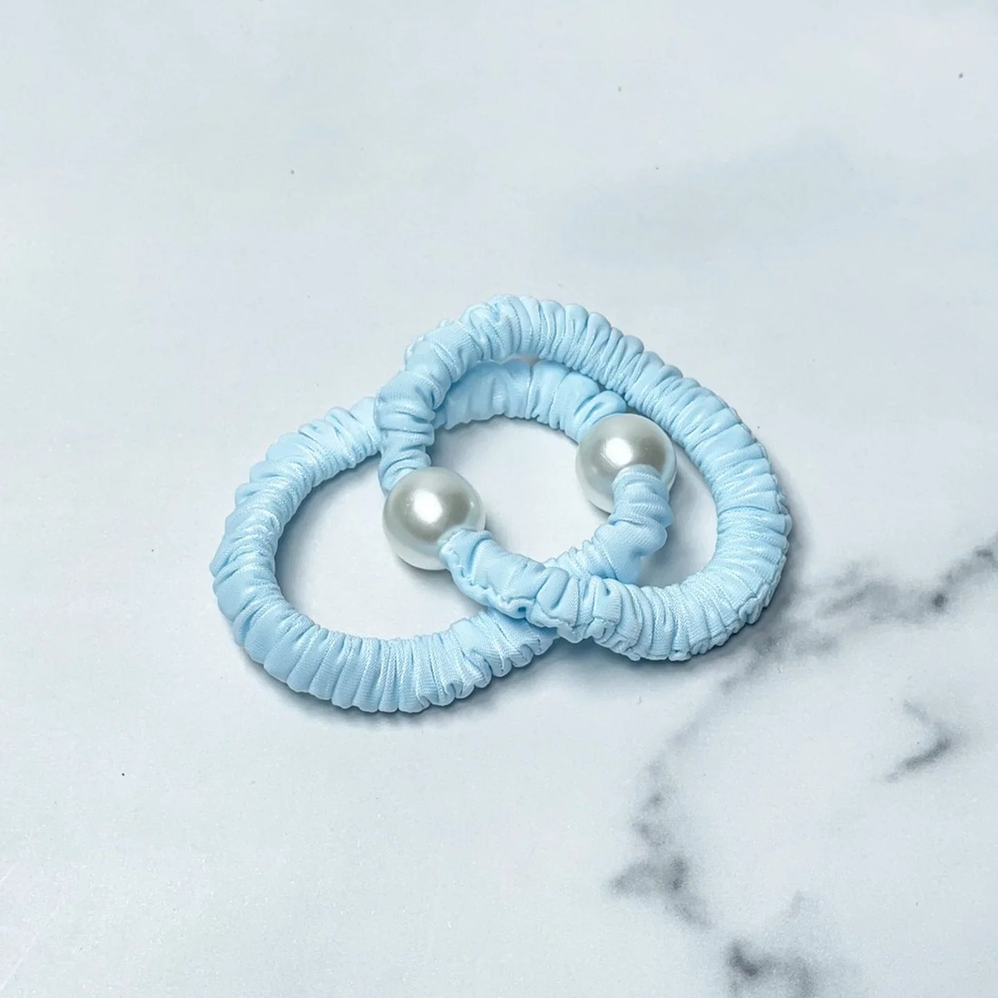 Two blue pastel hair ties with pearl detail on a white marble surface