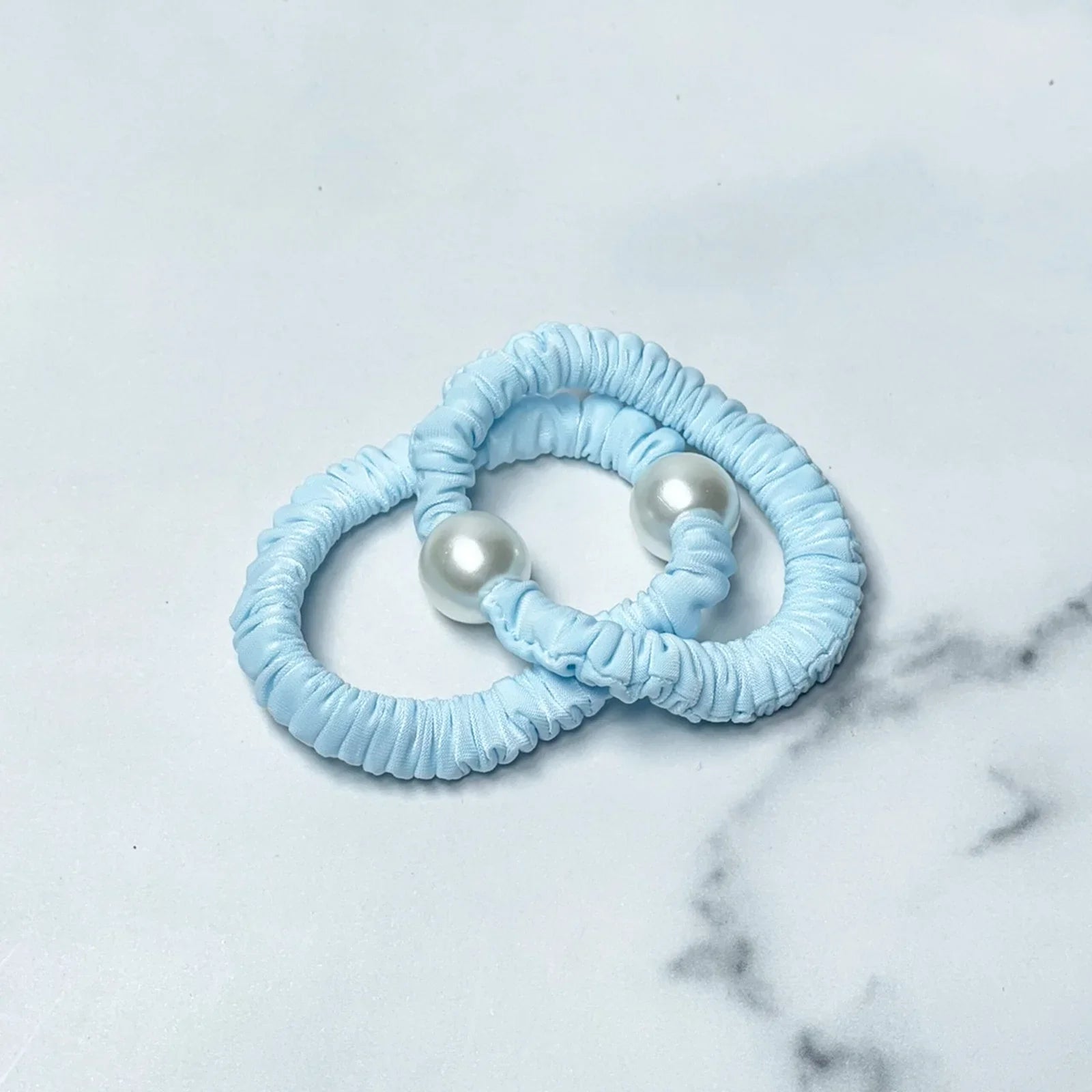 Two blue pastel hair ties with pearl detail on a white marble surface