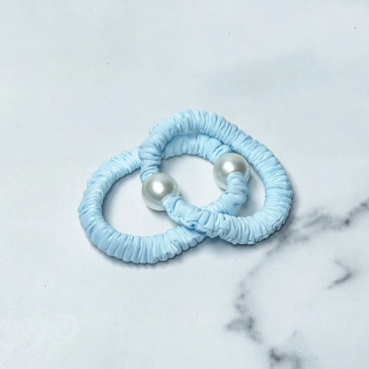 Two blue pastel hair ties with pearl detail on a white marble surface