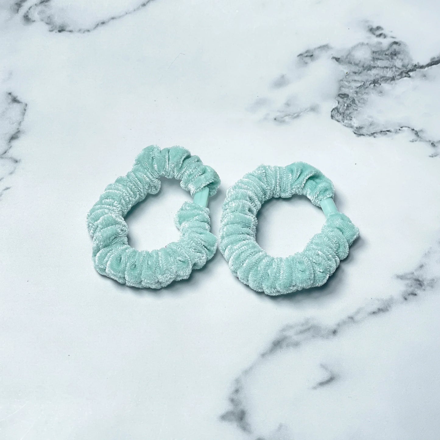 Two light blue shiny velvet hair ties on a white marble surface