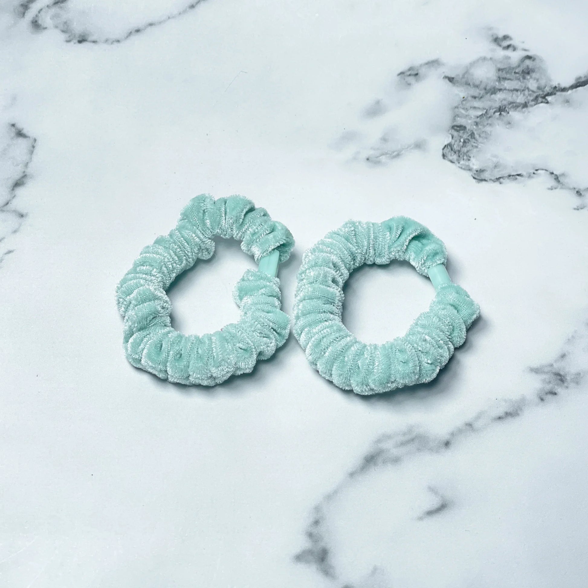 Two light blue shiny velvet hair ties on a white marble surface