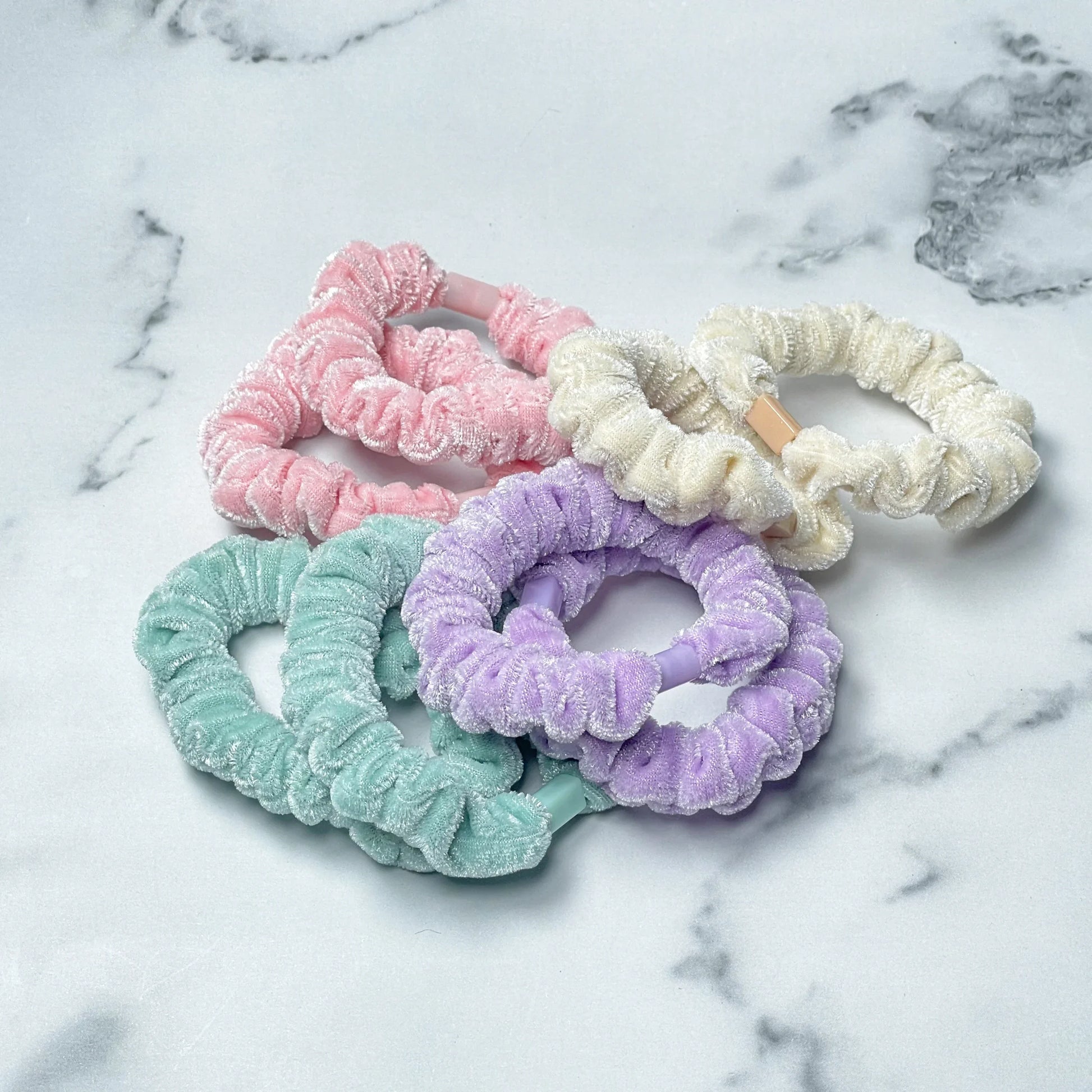 Set of shiny velvet hair ties in pastel pink, cream, purple, and teal colors on marble surface