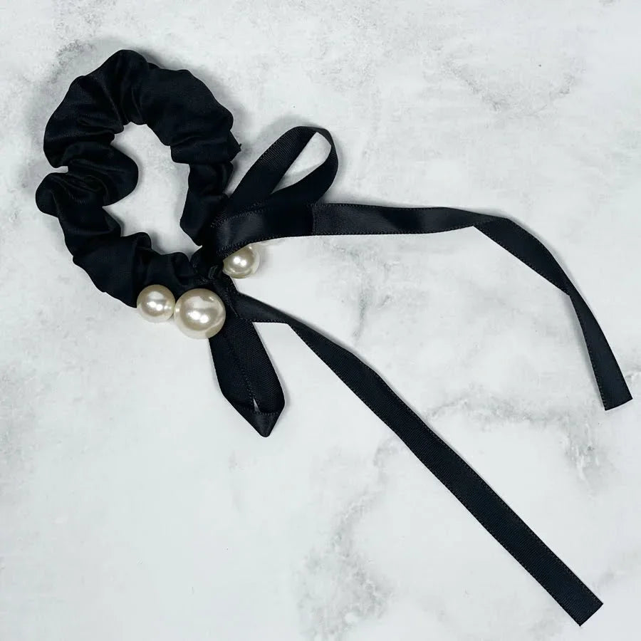 Black soft cloth hair scrunchie with decorative pearls and long ribbon tails on a white marble background