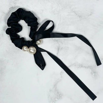 Black soft cloth hair scrunchie with decorative pearls and long ribbon tails on a white marble background